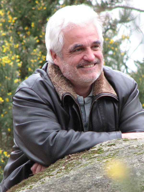 Portrait de Pierre Thirault, fondateur de l’Institut Pierre Thirault, souriant avec bienveillance dans un cadre naturel. Il porte une veste en cuir avec un col en fourrure, appuyé sur un rocher recouvert de mousse. En arrière-plan, des arbustes avec de petites fleurs jaunes ajoutent une touche lumineuse à l’image. Cette photo reflète sa personnalité chaleureuse et son engagement pour le bien-être et la connaissance de soi