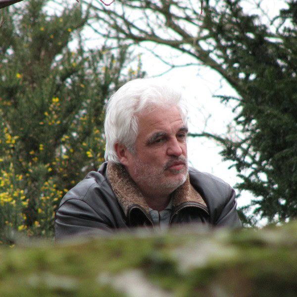 Portrait de Pierre Thirault, fondateur de l’Institut Pierre Thirault, capturé en extérieur dans un cadre naturel. Il porte une veste avec un col en fourrure et regarde au loin, entouré de verdure et de branches d’arbustes fleuris. L’image reflète une atmosphère de réflexion et de sérénité, en lien avec ses enseignements sur le bien-être, la géobiologie et la connaissance de soi