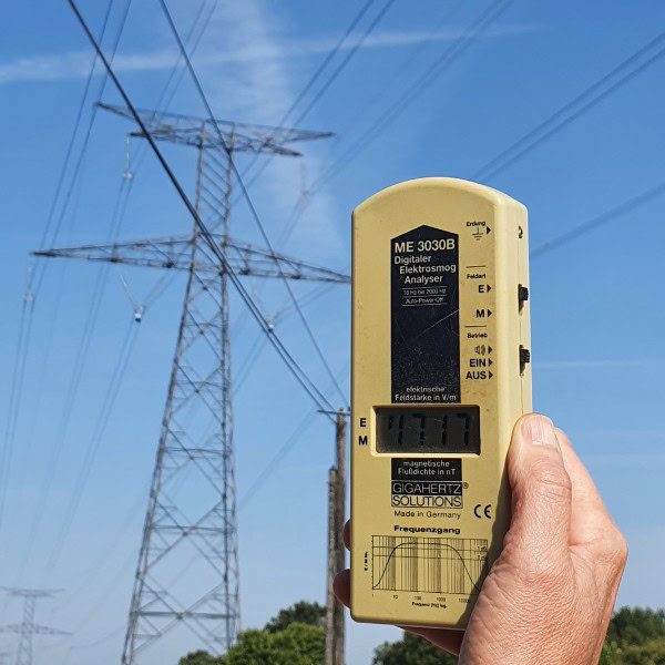 Un appareil de mesure des champs électromagnétiques, tenu par une main, est pointé vers une ligne à haute tension en arrière-plan. L’image illustre une analyse environnementale pour détecter les ondes électromagnétiques, en lien avec les pratiques de géobiologie et de bien-être de l’Institut Pierre Thirault